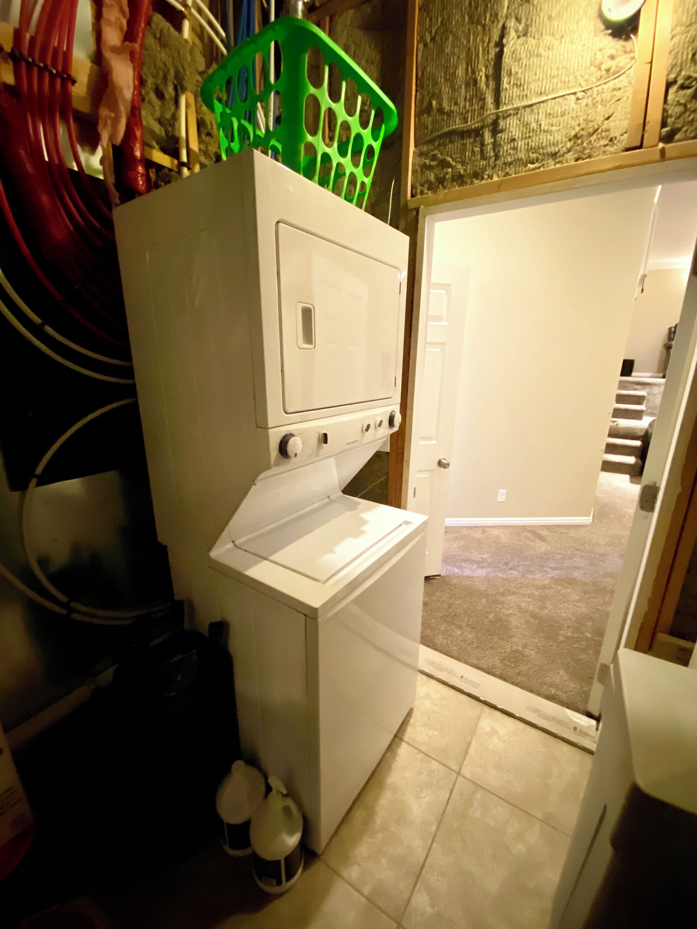 A compact laundry room with a stacked washer and dryer, a green laundry basket on top, and a door leading to a hallway.