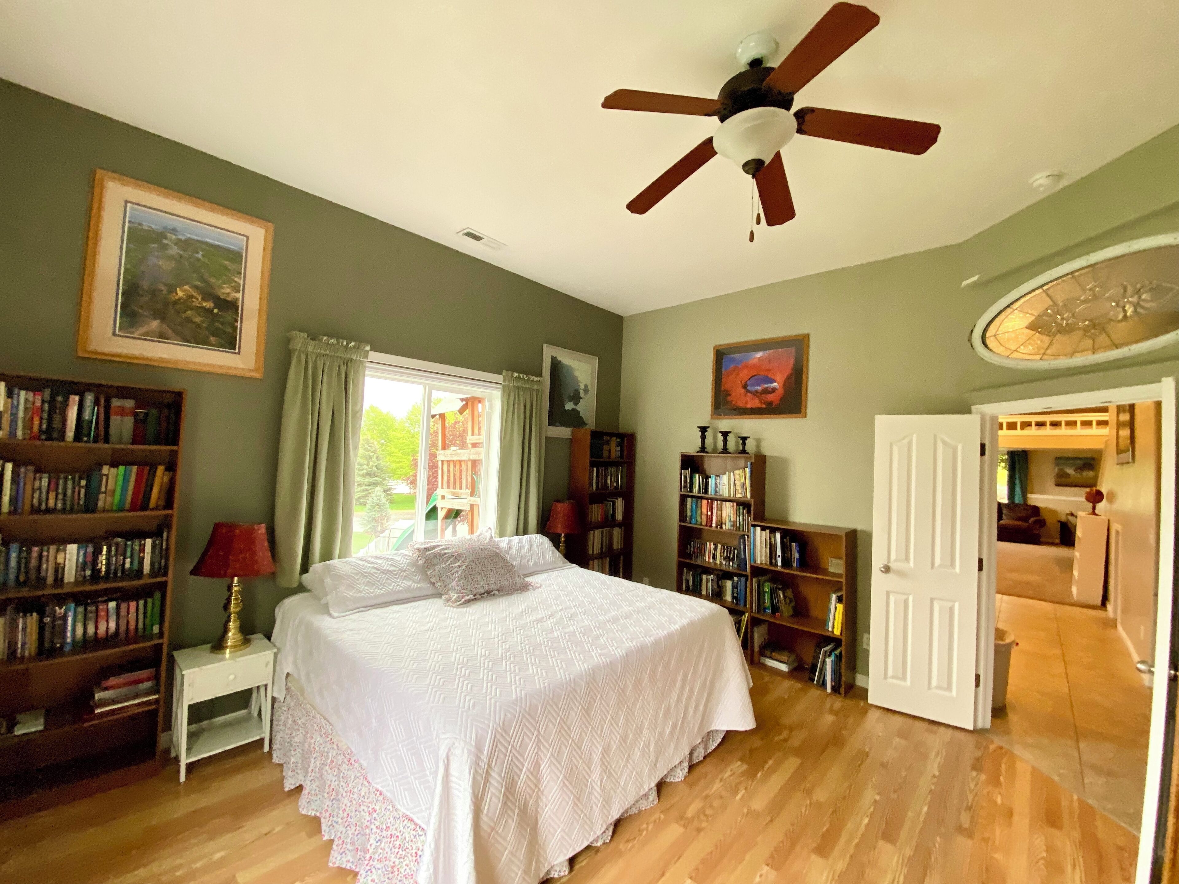 A bedroom with a bed, a ceiling fan, a bookshelf, and a door leading to another room.