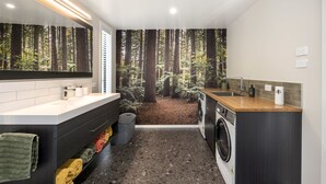 Un baño moderno con un espejo grande, un lavamanos, una lavadora y vistas a un bosque por una ventana.