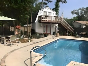 A swimming pool with a wooden deck, a covered patio area, and a two-story building with a balcony.