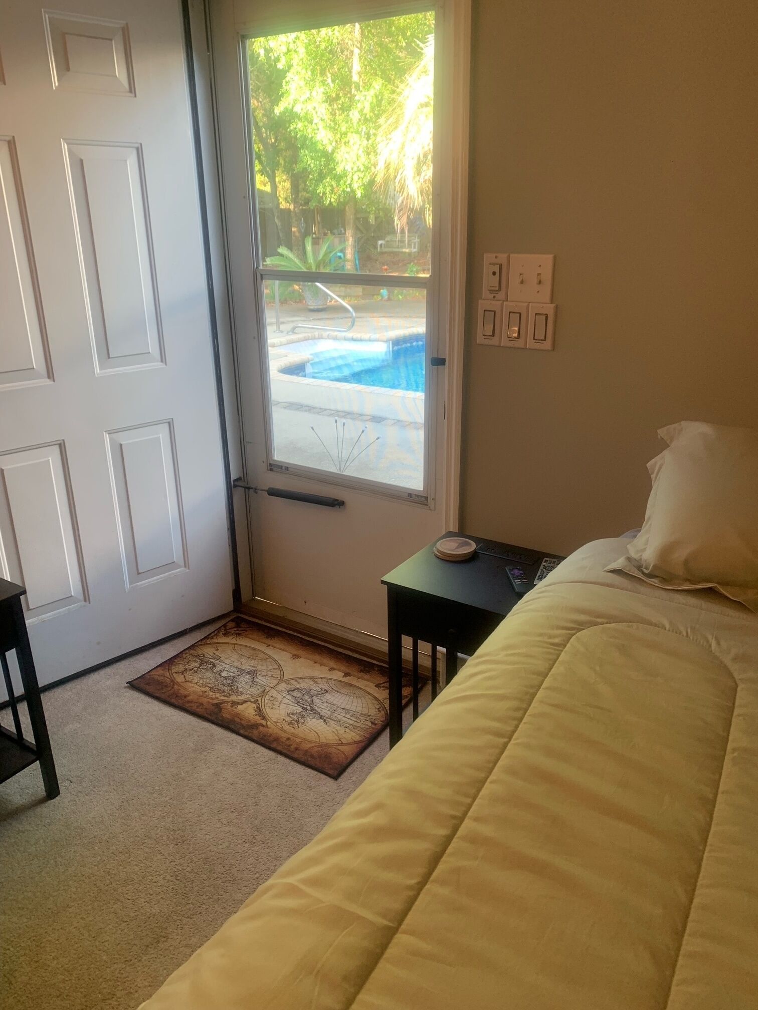 A hotel room with a bed, a small table, a window with a view of a swimming pool, and a door.