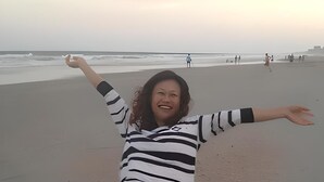 Eine Person sitzt am Strand, die Arme ausgebreitet, und trägt ein gestreiftes Hemd und Jeansshorts.