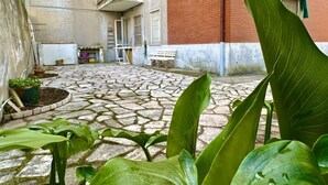 A courtyard with a stone floor, potted plants, a bench, and a building with red brick and white walls. - S325 - Sirolo, four-room apartment in the center with garden and parking space (SIROLO)