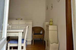 Une petite pièce avec une table blanche, une chaise en osier, un meuble et un radiateur.