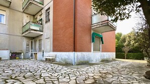 A courtyard with a stone floor, a building with a red brick facade, and a green awning over a window. - S325 - Sirolo, four-room apartment in the center with garden and parking space (SIROLO)