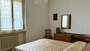 A bedroom with a bed, a dresser, a chair, a radiator, and a window with curtains. - S325 - Sirolo, four-room apartment in the center with garden and parking space (SIROLO)