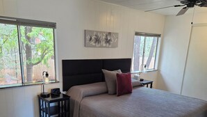 Une chambre à coucher avec un grand lit, une table de chevet avec une lampe, un ventilateur de plafond et une fenêtre donnant sur des arbres.