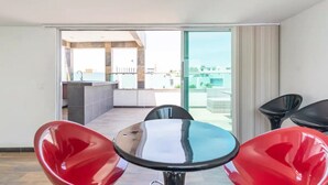 Una habitación moderna con un ventanal que da a un balcón. Hay una mesa redonda con sillas, una zona de cocina y vistas de la ciudad.
