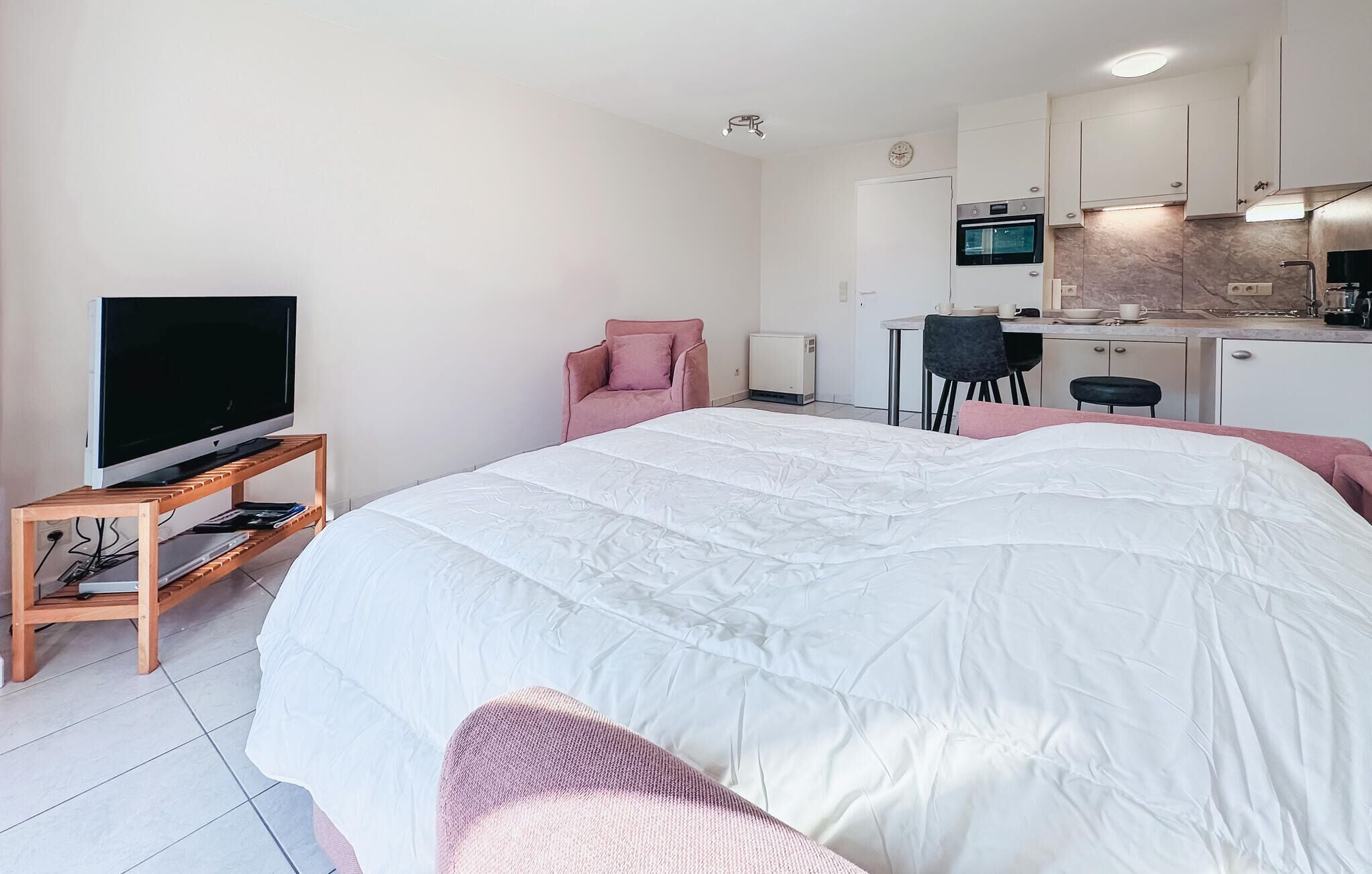Quarto de hotel moderno com uma cama grande, uma televisão em um suporte de madeira, uma cozinha compacta e uma área de estar com uma cadeira e uma mesinha.