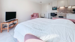 Una habitación de hotel moderna con una cama grande, un televisor sobre un soporte de madera, una kitchenette y un rincón de descanso con una silla y una mesita.