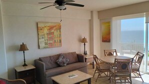 Una sala de estar con sofá, mesa de centro y comedor con sillas. Hay un ventilador de techo, dos lámparas y un cuadro en la pared.