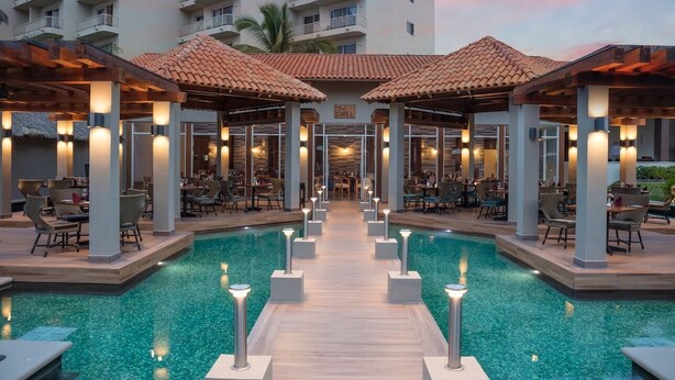 Zona de piscina con terraza de madera, asientos al aire libre y un restaurante con techo de tejas.