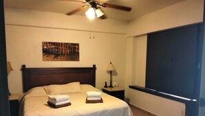 Una habitación de hotel con cama, mesita de noche, lámpara, ventilador de techo y un ventanal grande.