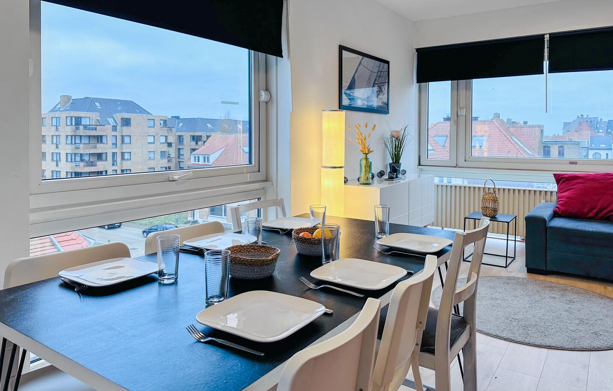 Ein Essbereich mit Tisch für vier Personen, großen Fenstern mit Blick auf die Stadt, einem roten Kissen auf einer dunklen Couch und einer Vase mit getrockneten Blumen auf einem Regal.