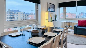 A dining area with a table set for four, large windows with city views, a red cushion on a dark sofa, and a vase with dried flowers on a shelf. - Nice apartment in Oostende with Wi-Fi (Oostende)