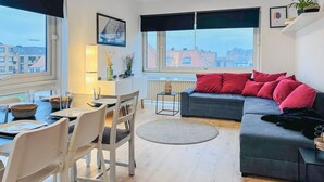 A modern living room with a grey sofa, red cushions, a dining table with chairs, and a view of buildings through large windows. - Nice apartment in Oostende with Wi-Fi (Oostende)