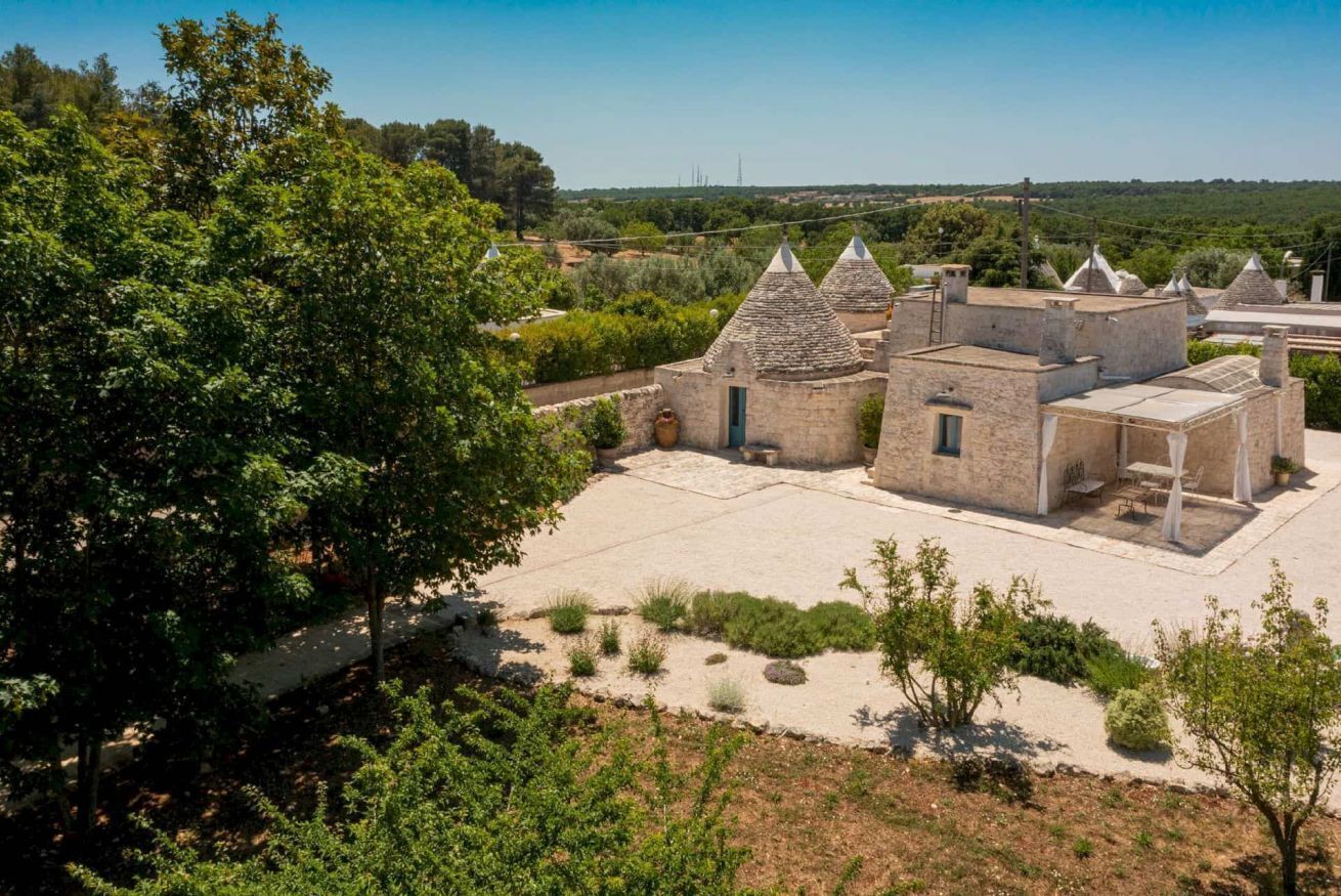 A traditional stone building with multiple conical roofs, surrounded by greenery and a clear sky.