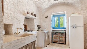 A rustic kitchen with stone walls, a gas stove, a sink, a white refrigerator, and a window with blue shutters.