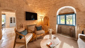 A stone-walled room with a sofa, a coffee table, a TV, and a window with blue shutters. - Trullo Pia - Trullo with private pool (Monopoli)