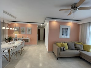 A modern living room with a beige sofa, a dining area with a white table and chairs, and a kitchen in the background.