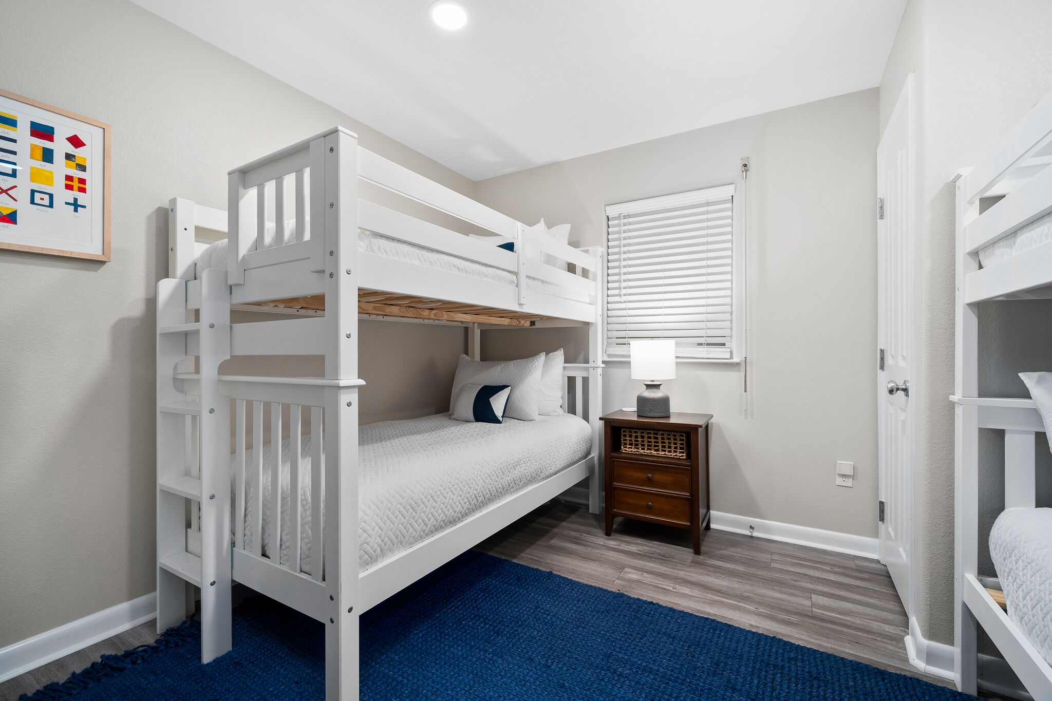 Habitación con literas, una alfombra azul, una mesita de noche con lámpara y una ventana con persianas.