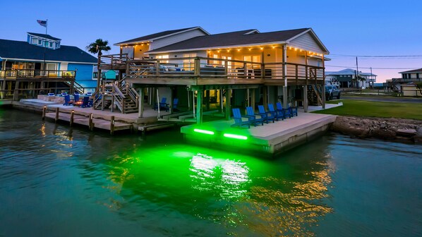 Ein Haus am Wasser mit Terrasse, Sitzgelegenheiten im Freien und einem Pool mit grüner Beleuchtung.