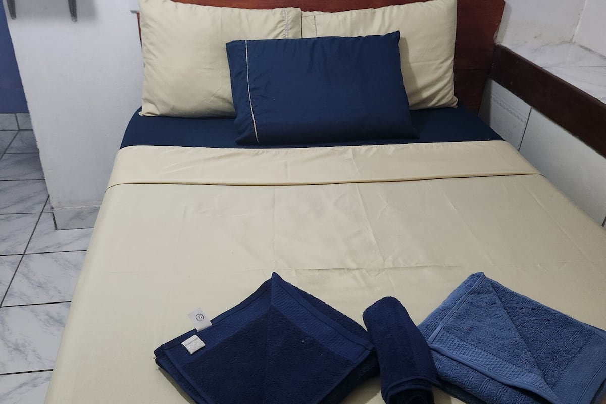 A neatly made bed with a beige bedspread and blue pillows, a wooden headboard, and a small table to the left.
