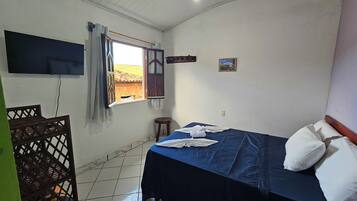 A bedroom with a bed, a TV mounted on the wall, a window with curtains, and a small stool.
