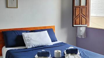 Un lit bien fait, avec une literie bleue et blanche, une tête de lit en bois et une fenêtre donnant sur une clôture blanche.