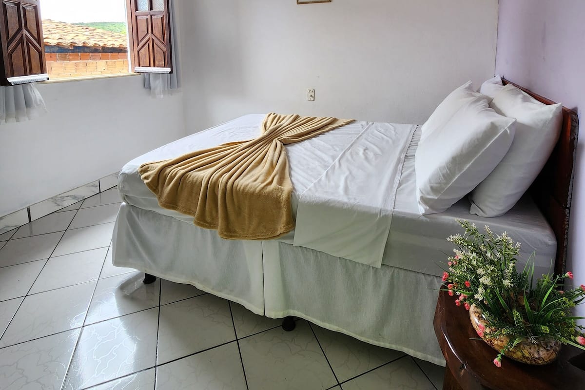 A bedroom with a bed, a ceiling fan, a window with curtains, a small framed picture on the wall, and a potted plant on a wooden table.