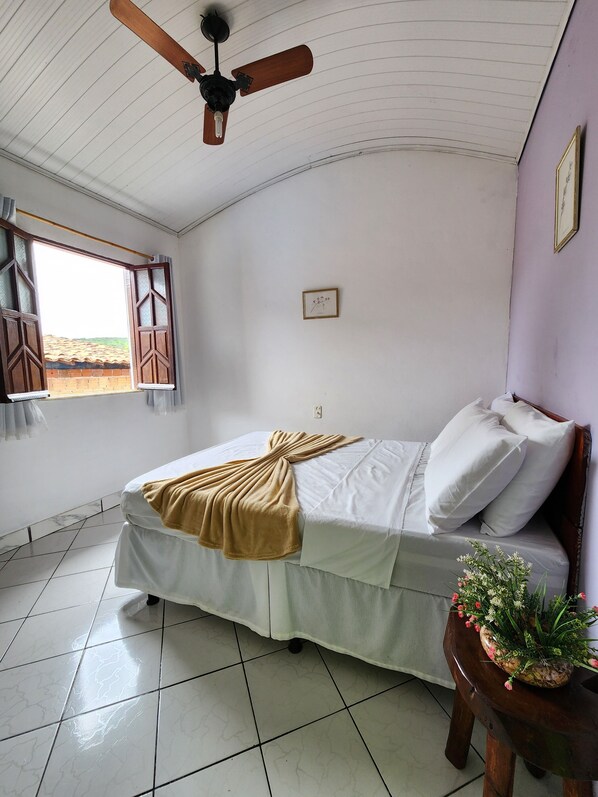 Un dormitorio con cama, ventilador de techo, ventana con cortinas, un pequeño cuadro enmarcado en la pared y una planta en maceta sobre una mesa de madera.