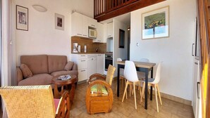 En kompakt opholdsstue med sofa, spisebord, stole, et lille køkken og en trappe.