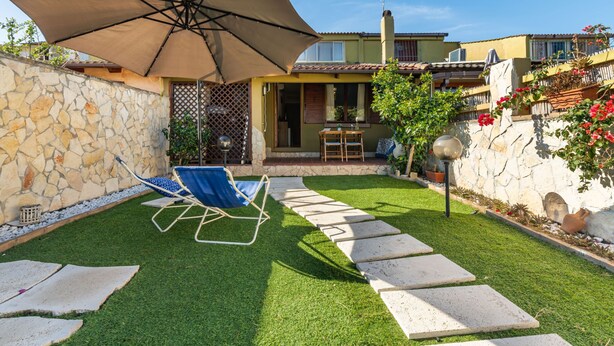 Un jardin avec un mur en pierre, une pelouse, une terrasse avec une table et des chaises, et un grand parasol.
