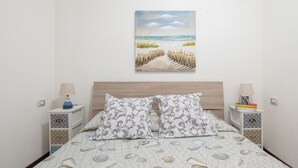 Une chambre à coucher avec un lit, deux tables de chevet, un tableau représentant une scène de plage et une literie à motifs.