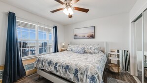 A bedroom with a bed, a ceiling fan, a window with curtains, a nightstand, a bedside table, and a painting on the wall. - Sunsets Over Lake Padre | Canal Home | 2 blk to beach (Corpus Christi)