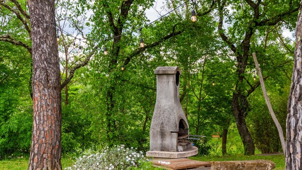 Un cadre extérieur rustique, avec une structure en pierre, peut-être une cheminée, entouré d’une végétation luxuriante et d’arbres.
