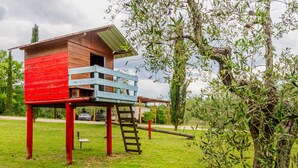 Une maison de jeu en bois, avec une façade rouge et des touches de bleu, située sur une pelouse, avec des arbres en arrière-plan.