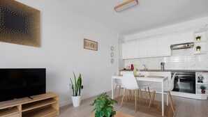 Een moderne woonkamer met een flatscreen-tv, een eethoek met een witte tafel en stoelen, en een keuken met witte kasten en een inbouwoven.