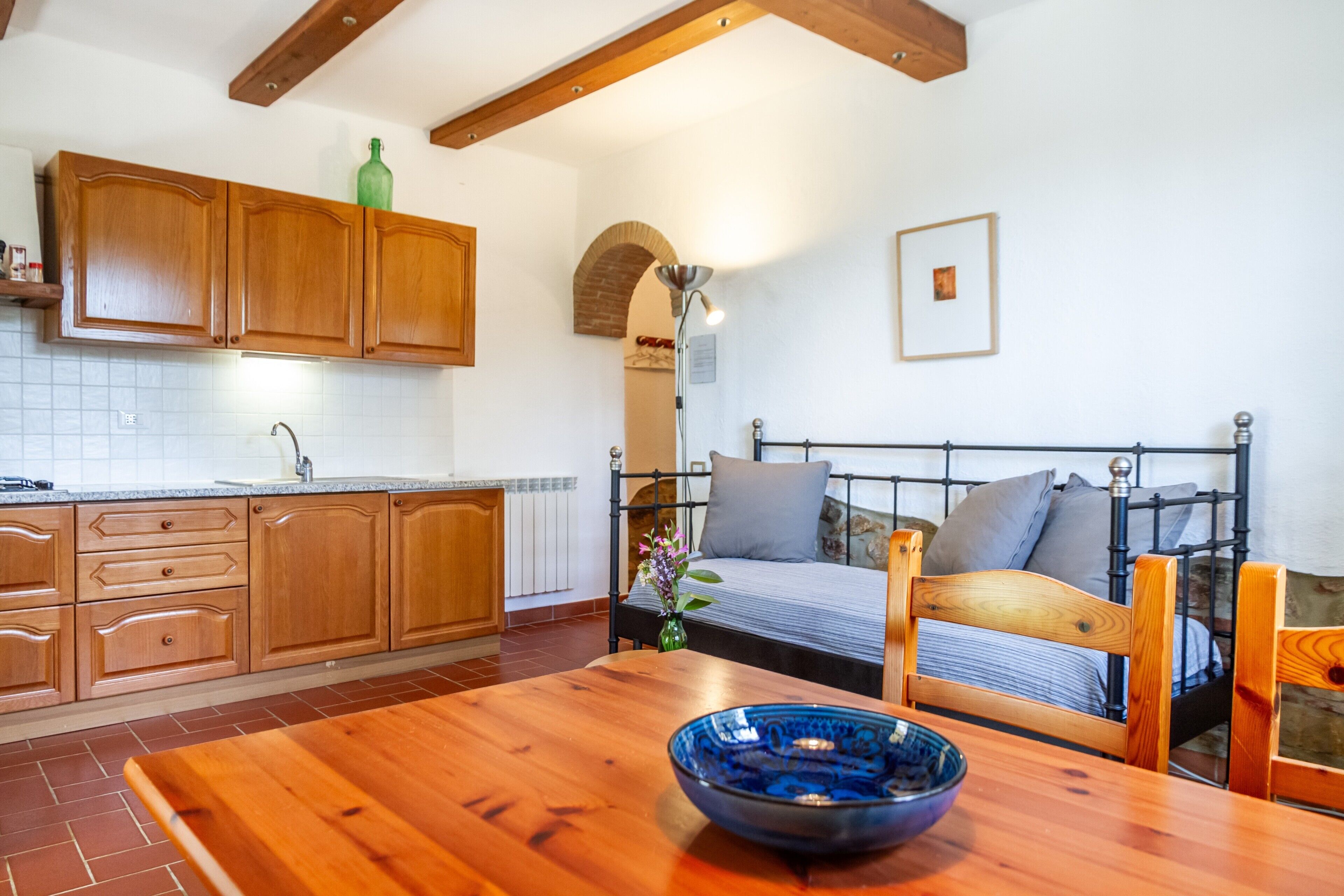 Une cuisine avec des meubles de rangement en bois, une table à manger sur laquelle se trouve un saladier bleu, et un lit avec un cadre en métal.