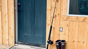 En træhytte med en mørkeblå dør, et fiskestang lænet op ad væggen, og et par støvler på verandaen.
