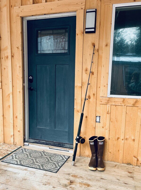 A wooden cabin with a dark blue door, a fishing rod leaning against the wall, and a pair of boots on the porch.