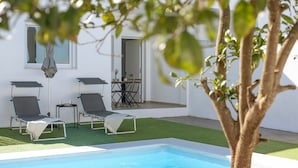 Un área de piscina con sillones de descanso y una mesa, rodeada de paredes blancas y un árbol.