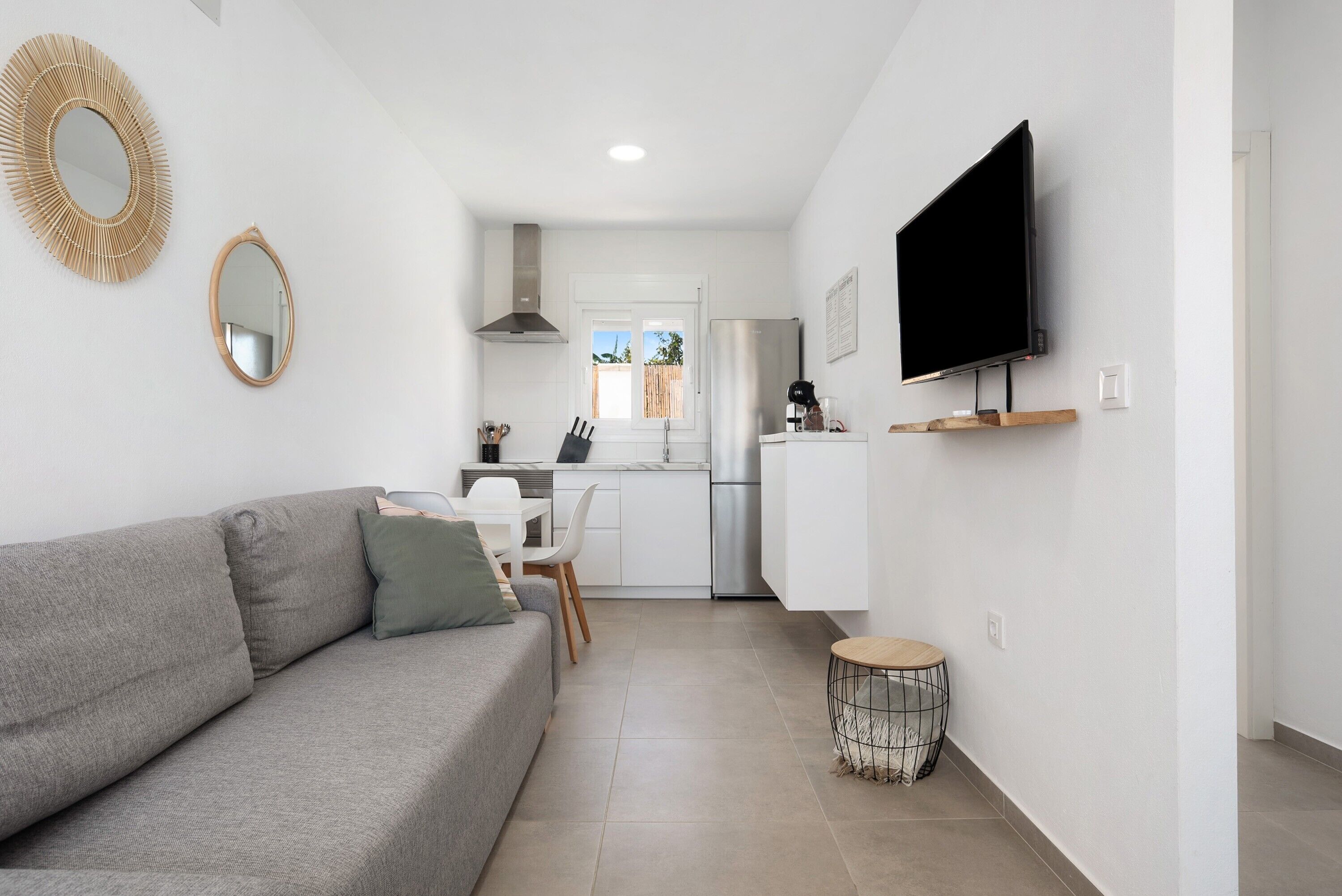 Un salon moderne avec un canapé gris, un téléviseur à écran plat fixé au mur et une cuisine en arrière-plan.