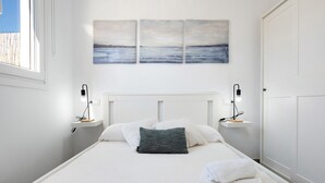 Una cama bien hecha con ropa blanca y una sola almohada gris, flanqueada por dos mesitas de noche con lámparas, en una habitación con pared blanca y vistas al mar a través de una ventana.