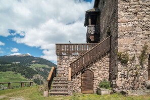 A stone building with a wooden staircase and balcony, surrounded by a mountainous landscape.