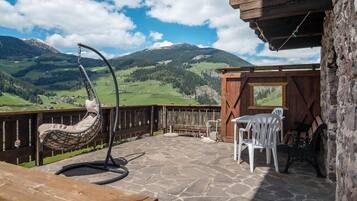 Une terrasse en bois avec un fauteuil suspendu, une table et des chaises, offrant une vue sur un paysage montagneux.