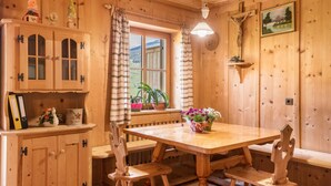 Un intérieur en bois avec une table en bois, des bancs, une fenêtre avec des rideaux, une étagère fixée au mur avec un crucifix et un tableau encadré.