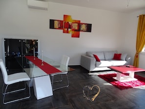 A modern living room with a glass dining table, white chairs, a grey sofa with red cushions, and abstract wall art.