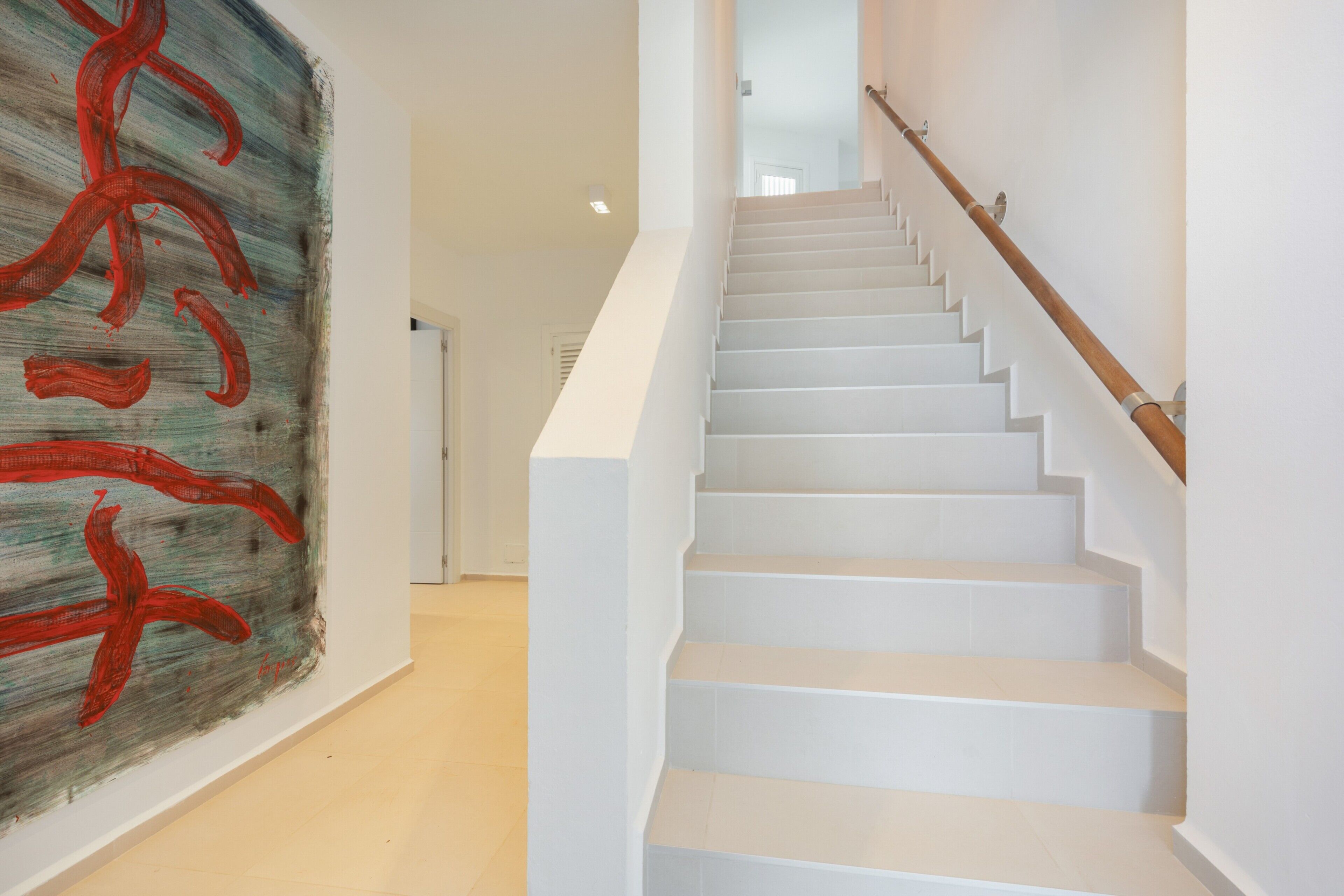 Una escalera con escalones blancos y barandilla de madera que conduce a una habitación con un cuadro abstracto grande en la pared.