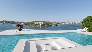 Une piscine au design géométrique, des chaises longues blanches et une vue sur une ville côtière.